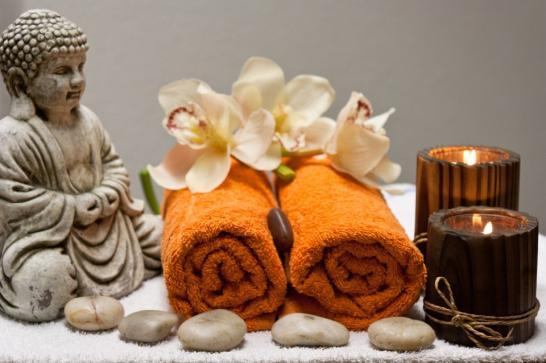 Mise en scène de bien-être avec un petit bouddha, 2 essuies oranges roulés, 2 fleurs par-dessus, 2 bougies brunes allumées et quelques galets déposés.