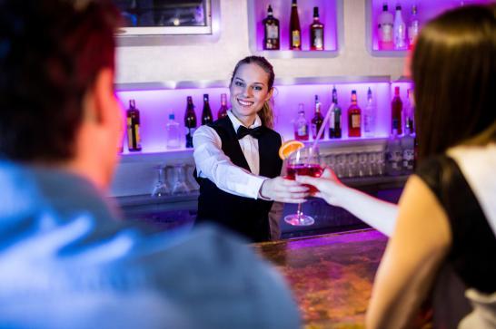 Barmaid en costume cravate qui tend un verre de cocktail à une cliente de l'autre côté du bar