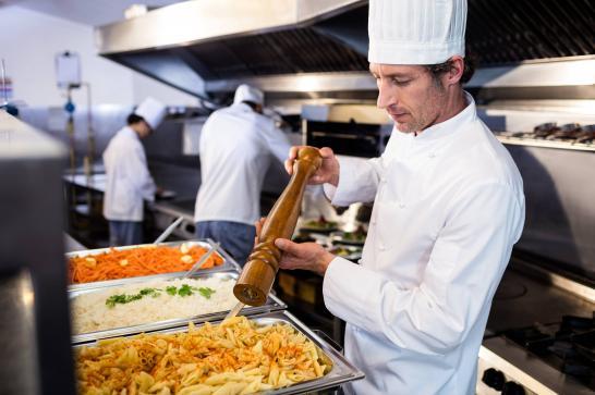 Cuisinier dans une cuisine qui mout du poivre au-dessus d'une très grand plat de pâtes, il y a aussi un gros plat de riz et de crudités