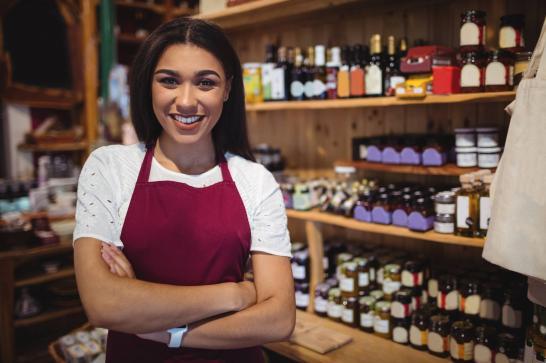 Gérante d'un point de vente vêtue d'un tablier de travail qui regarde l'appareil photo. Elle se trouve dans son comptoir, devant une étagère remplie de produits