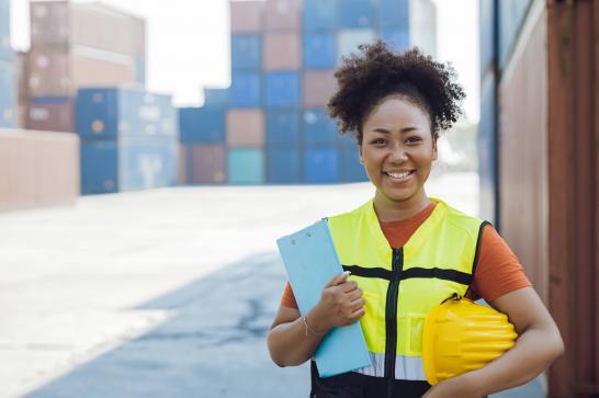 Déclarante en douane avec ses équipements de sécurité et son portefolio de documents en main. Elle se situe dans un port industrielle et est entourée de containers