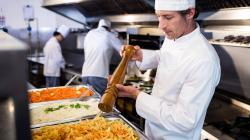 Cuisinier dans une cuisine qui mout du poivre au-dessus d'une très grand plat de pâtes, il y a aussi un gros plat de riz et de crudités