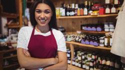 Gérante d'un point de vente vêtue d'un tablier de travail qui regarde l'appareil photo. Elle se trouve dans son comptoir, devant une étagère remplie de produits
